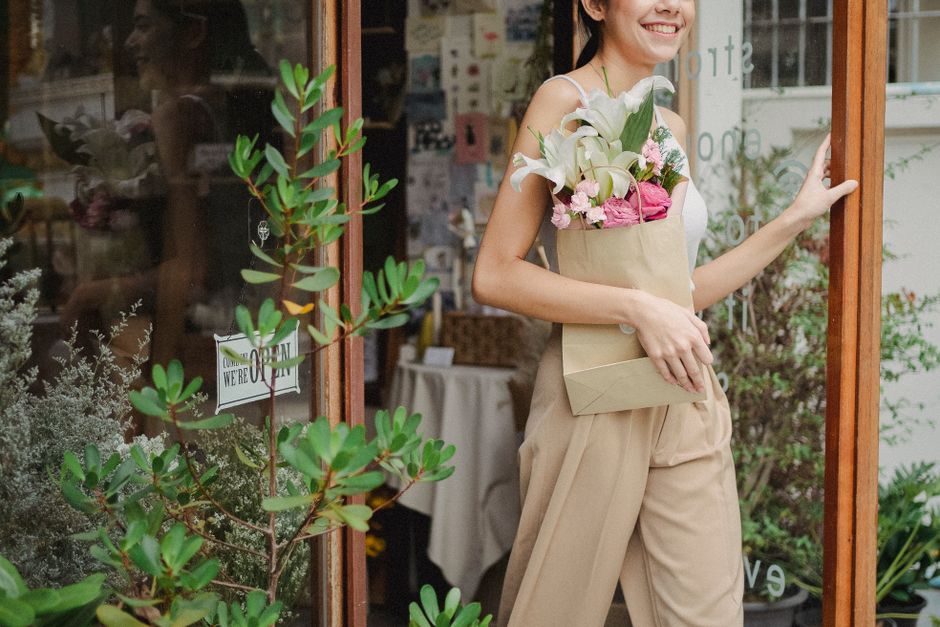 A woman with a cheerful expression holds a bouquet of fresh flowers, including white calla lilies and pink roses, wrapped in brown paper. She stands outside a floral shop, Cheam Flowers, with a glass 