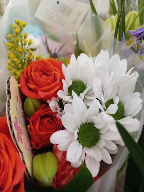 A close-up view of a floral arrangement featuring vibrant orange roses with tightly curled petals, complemented by white daisies with large, rounded petals and prominent green centers. The bouquet inc