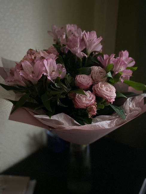 A bouquet of fresh pink flowers arranged with a mix of pale pink roses, alstroemeria, and lisianthus, complemented by dark green foliage. The flowers are wrapped in light pink tissue paper with a soft