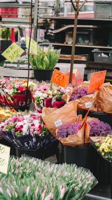 A vibrant display of fresh floral arrangements at an outdoor market stand, featuring various bouquet arrangements wrapped in brown paper and colorful cellophane. Prominent flowers include pink and whi