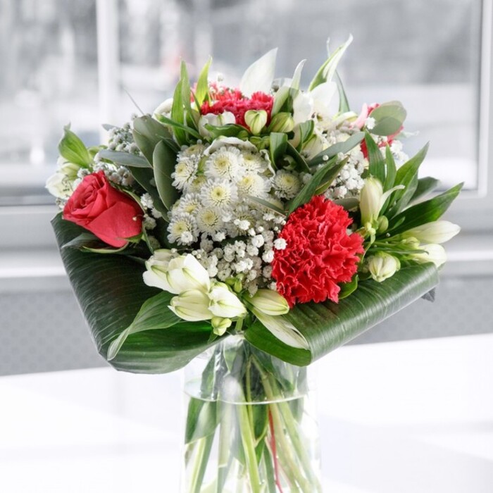 In this image, a luxurious red and white bouquet commands attention from the centre of a pristine white tabletop, set against a softly diffused, light-filled background that evokes a calm Cheam morning. The flowers are gathered in a tall, cylindrical clear glass vase, its sides catching subtle reflections while showcasing the straight, meticulously trimmed green stems beneath the waterline. At the heart of the arrangement, deep red roses unfurl their velvety petals, each bloom full and romantic, suggesting timeless affection. A large crimson carnation, intensely ruffled and layered, nestles towards the front, bringing drama and richness in both texture and colour. Threaded among these scarlet tones are cool white blossoms: slender-petalled alstroemeria with fine speckles near each throat, compact white chrysanthemums or gerbera-style blooms adding round softness, and several ivory lily buds still closed, hinting at future fragrance and longevity. Sprays of gypsophila, or baby's breath, scatter tiny white dots through the bouquet like stardust, lending it a light, airy feel that softens the strong reds. Surrounding this blend of flowers is an abundance of greenery, carefully arranged to frame and lift the composition. Broad, glossy dark green leaves are artfully wrapped and curled around the base, forming a structured collar that grounds the bouquet, while eucalyptus-like sprigs and pointed green foliage rise between the stems, adding different shades and shapes of green for depth. The overall mood is polished yet welcoming, like a centrepiece set out for a family celebration in a Cheam home after a stroll through Nonsuch Park. Crafted with the quiet confidence of an experienced local florist, this bouquet would feel equally at home marking an anniversary, congratulating a new achievement, or bringing brightness to a Cheam office desk with same-day delivery.